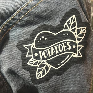 May include: A black and white heart-shaped patch with a banner that reads "POTATOES." The patch is sewn onto a blue denim fabric.