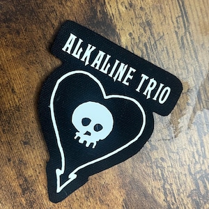Könnte beinhalten: Schwarzer Aufnäher mit einem weißen herzförmigen Design mit einem Totenkopf und dem Text "ALKALINE TRIO" in Weiß. Der Aufnäher hat eine einzigartige Form mit spitzem Boden.