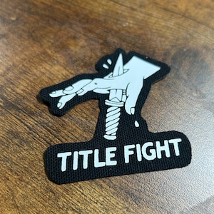 Puede incluir: Parche bordado en blanco y negro con una mano sosteniendo un cuchillo y el texto "TITLE FIGHT".