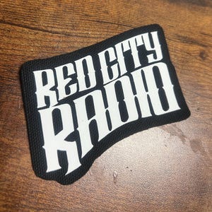 May include: Black rectangular patch with the words "RED CITY RADIO" in bold white letters. The patch has a black border and is set against a brown wooden surface.