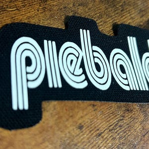 May include: Black patch with the word "piebald" in white, stylized, retro font. The patch has a unique shape with a notch on one side. The background is a wooden surface.