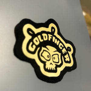 Peut inclure: Patch de couleur or avec un crâne et le texte "Goldfinger" sur un fond noir.