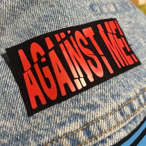 May include: A black patch with the words "AGAINST ME!" in red letters sewn onto a light blue denim fabric.