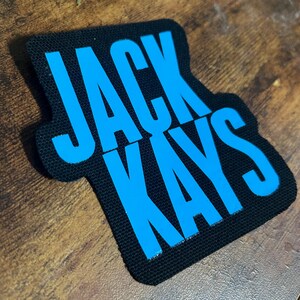 May include: A black fabric patch with the words "JACK KAYS" in blue lettering.