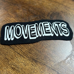 Op de afbeelding: Zwarte stoffen patch met witte tekst "MOVEMENTS".