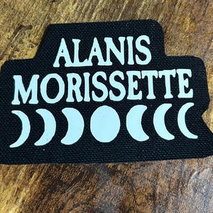 May include: Black fabric patch with white text that reads "Alanis Morissette" and five white crescent moons.