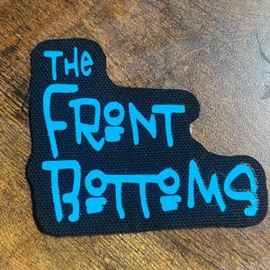 Peut inclure: Écusson en tissu noir avec les mots "The Front Bottoms" écrits en bleu.