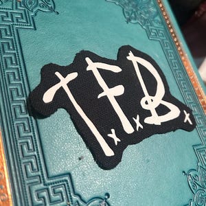 May include: Black fabric patch with white lettering that reads "TFB" with two white "x" marks below the letters.