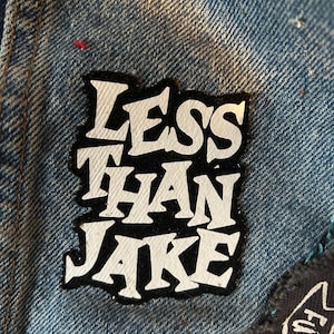 Op de afbeelding: Close-up van een spijkerjasje met een patch met de tekst "LESS THAN JAKE" in witte blokletters op een zwarte achtergrond. De jas heeft een lichtblauwe wassing.