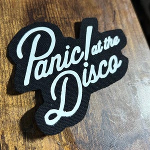 Könnte beinhalten: Schwarzer Stoff-Patch mit weißer Aufschrift "Panic! at the Disco" in kursiv.