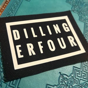 Könnte beinhalten: Schwarzer Stoff-Patch mit weißem Text, der "DILLING ERFOUR" lautet.