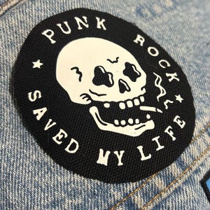 Pode incluir: Patch bordado preto e branco com uma caveira fumando um cigarro. O texto em torno da caveira diz "Punk Rock Saved My Life".