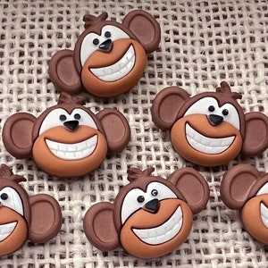 May include: Six brown monkey face buttons with white teeth and black eyes. The buttons are on a brown burlap background.