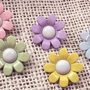 May include: Five pastel colored daisy shaped buttons with white centers. The buttons are arranged in a semi-circle on a beige burlap background.
