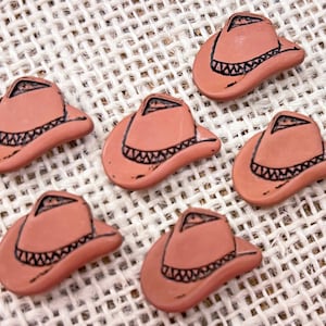 May include: Six brown cowboy hat shaped buttons with black trim. The buttons are on a beige woven fabric background.