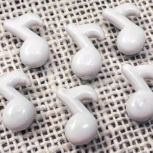 May include: Six white ceramic eighth note charms. Each charm has a glossy finish and a rounded shape. The charms are arranged on a textured, neutral-colored surface, suggesting a craft or jewelry-making theme.