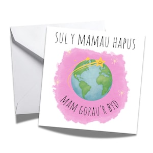 Puede incluir: Una tarjeta de felicitación blanca con un fondo rosa de acuarela y las palabras "SUL Y MAMAU HAPUS" y "MAM GORAU'R BYD" en negro. La tarjeta presenta un globo terráqueo con estrellas fugaces y una estrella sonriente.