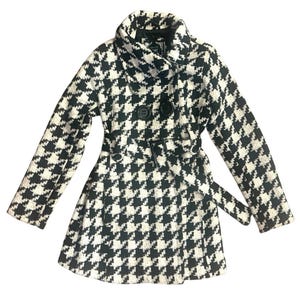 May include: A black and white houndstooth patterned coat featuring a wide collar, long sleeves, and a double-breasted front with two large black buttons. The coat includes a matching fabric belt with a black buckle at the waist.
