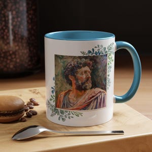 May include: White ceramic mug with a teal rim and handle. The mug features a watercolor painting of a man's face with a beard, surrounded by green leaves.
