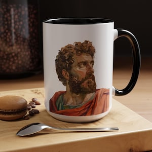 May include: White ceramic mug with a black rim and handle. The mug features a colorful illustration of a man with a beard and curly hair, wearing a red and gold robe.
