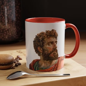 May include: White ceramic mug with a red interior and handle. The mug features a portrait of a man with a beard and curly hair. A spoon and a macaron are on a wooden surface next to the mug.