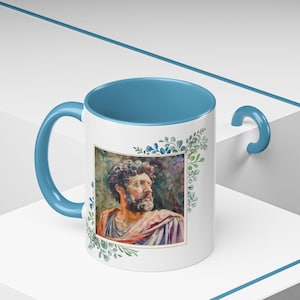 May include: A white ceramic mug with a blue interior and handle. The mug features a watercolor illustration of a man's face with a beard, surrounded by green leaves. The image is framed with a gold border.