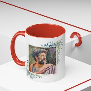 May include: A white ceramic mug with a red rim and handle. The mug features a watercolor illustration of a man with a beard, surrounded by green and blue leaves.