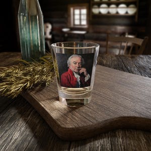 May include: A clear glass shot glass featuring a printed image of a man in a red jacket and white shirt. The man is looking to the side and has his hand on his chin.