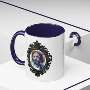 May include: White ceramic mug with a blue rim and handle. The mug features a watercolor portrait of a man with a beard, framed in an ornate gold design.