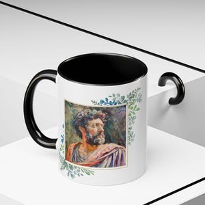 May include: A black and white mug with a black handle. The mug has a white design of a man's face with a beard and a floral border around the image.