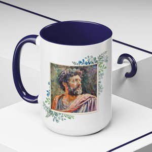 May include: White ceramic mug with a navy blue handle and rim. The mug features a watercolor painting of a man's face with a beard, surrounded by a green and blue floral border. The mug is a great gift for art lovers.