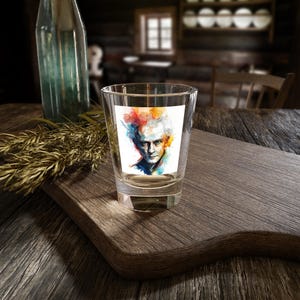 May include: A clear glass shot glass featuring a colourful watercolour portrait of a man with grey hair. The portrait is printed on a white sticker and is centred on the glass.