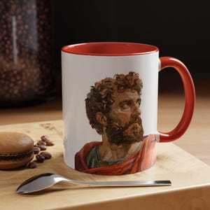 May include: A white ceramic mug with a red rim and handle. The mug features a colorful illustration of a man with a beard and curly hair. The man is wearing a red and green robe.