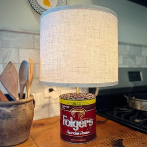 Puede incluir: Una lámpara hecha de una lata de café Folgers llena de granos de café. La lámpara tiene una pantalla de lino blanca con un ribete gris. La lata de café es roja y tiene el texto "Folgers Mountain Grown Coffee Special Roast" impreso en ella.