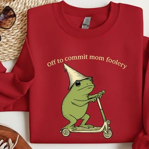 May include: A red sweatshirt with the phrase "Off to commit mom foolery" in a serif font. The graphic features a green frog wearing a pointed hat and riding a scooter. The sweatshirt has long sleeves.