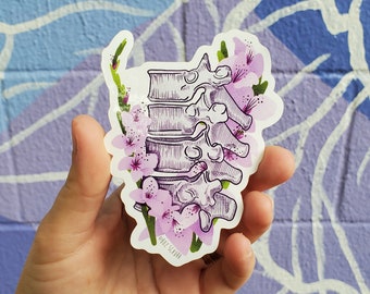 Könnte beinhalten: Ein Aufkleber mit einer detaillierten Illustration einer Wirbelsäule, umgeben von zarten rosa Kirschblüten. Die Wirbelsäule ist in Grautönen gehalten, während die Blüten einen Farbtupfer setzen. Der Aufkleber hat einen weißen Rand.