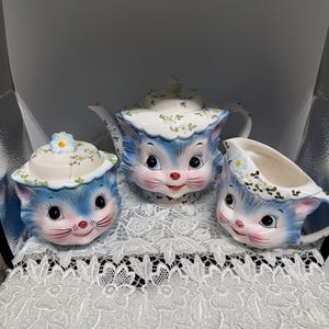 May include: A ceramic tea set featuring a teapot, a lidded sugar bowl, and a creamer, each shaped like a cat head with blue fur, large dark eyes, and pink noses. The lids and rims are decorated with small green leaves and tiny blue flowers.