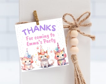 Könnte beinhalten: Eine weiße Karte mit dem Text "THANKS For coming to Emma's Party" wird von einer goldenen Klammer gehalten. Die Karte zeigt drei Aquarellkätzchen mit Partyhüten. Eine Schnurschleife und Holzperlen sind angebracht.