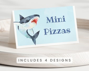 May include: A blue and white shark illustration with an open mouth and sharp teeth on a light blue background. The text "Mini Pizzas" is written in dark blue letters below the shark. The text "INCLUDES 4 DESIGNS" is written in black letters below the image.