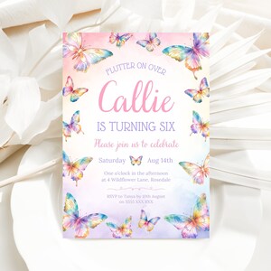 Puede incluir: Una invitaci&oacute;n de cumplea&ntilde;os rosa y morada con tem&aacute;tica de mariposas para una ni&ntilde;a que cumple seis a&ntilde;os. La invitaci&oacute;n presenta un dise&ntilde;o de acuarela con mariposas coloridas y el texto "Flutter on over Callie is turning six. Please join us to celebrate Saturday Aug 14th One o'clock in the afternoon at 4 Wildflower Lane, Rosedale RSVP to Tanya by 10th August on 5555 XXX XXX"
