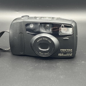 May include: A black Pentax IQZoom 80-E camera with a zoom lens, 38mm-80mm. The camera has a built-in flash and a wrist strap. The camera is on a dark surface.