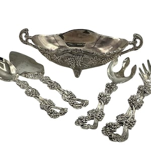 Puede incluir: Plato de servir de plata con diseño de vid y dos utensilios de servir a juego. El plato tiene un borde festoneado y una base decorativa. Los utensilios son una cuchara y una cuchara ranurada.