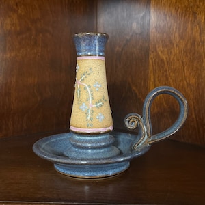 May include: A ceramic candle holder with a blue speckled base and handle. The central column features a tan speckled finish decorated with a painted floral vine pattern in green and light blue, accented by thin pink horizontal bands.