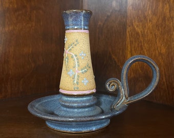 Könnte beinhalten: Ein Kerzenhalter aus Keramik mit blau gesprenkeltem Fuß und Henkel. Die mittlere Säule hat eine beige gesprenkelte Oberfläche, die mit einem gemalten Blumenrankenmuster in Grün und Hellblau sowie dünnen rosa Querstreifen verziert ist.