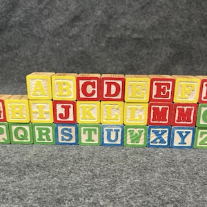 May include: A collection of wooden alphabet blocks arranged in three horizontal rows. Each cube features a letter of the alphabet in a bold, serif font. The letters are printed in various colors including red, yellow, blue, and green on natural wood.