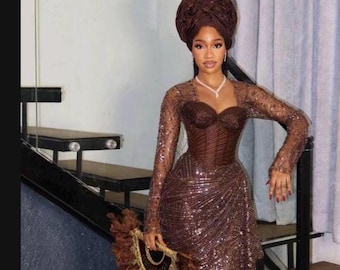 May include: A dark brown floor-length gown featuring long sleeves, a sweetheart neckline, and an all-over sequined pattern. The ensemble includes a matching structured headwrap and a decorative hand fan adorned with brown feathers and gold trim.