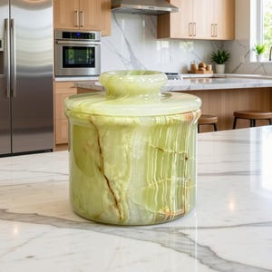 May include: A cylindrical light green stone canister with a matching lid. The stone features natural translucent banding and deep brown veining throughout the polished surface. The canister has a wide, flat base and a rounded, raised handle on the lid.