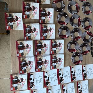 Puede incluir: Una colección de CD y estuches de CD. Los estuches de CD presentan el texto "The Elliotts" y un gráfico de un coche. Los CD tienen un diseño similar. La combinación de colores incluye rojo, blanco y negro.