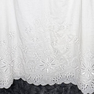 May include: White fabric with intricate embroidered details. The design features a dove, stars, and floral patterns along the hem. The fabric has a scalloped edge, adding a delicate touch to the overall aesthetic.