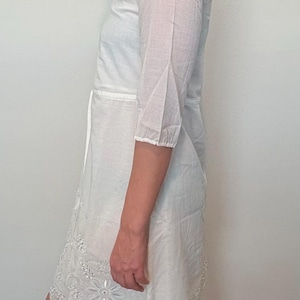May include: White eyelet dress with three-quarter sleeves and a tie waist. The dress features a floral embroidered hemline. The fabric appears to be lightweight and suitable for warm weather. The dress is knee-length.